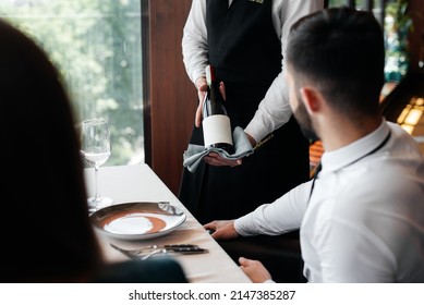 A Young Sommelier In A Stylish Apron Demonstrates And Offers Fine Wine To A Beautiful Couple In A Restaurant. Customer Service In An Elite Restaurant And A Public Restaurant.