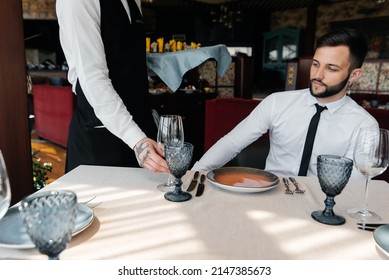 A Young Sommelier In A Stylish Apron Demonstrates And Offers Fine Wine To A Beautiful Couple In A Restaurant. Customer Service In An Elite Restaurant And A Public Restaurant.