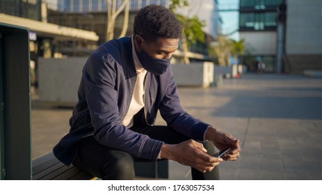 Young South African male with face mask holding mobile phone horizontally while sitting on city bench - Shutterstock ID 1763057534