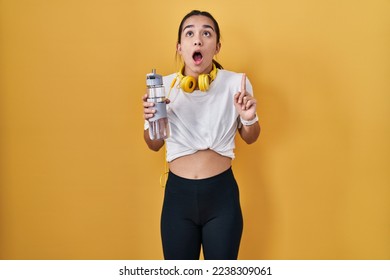 Young south asian woman wearing sportswear drinking water amazed and surprised looking up and pointing with fingers and raised arms.  - Shutterstock ID 2238309061