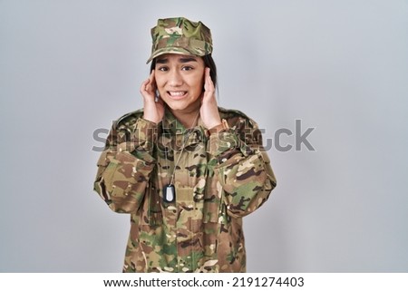 Young south asian woman wearing camouflage army uniform covering ears with fingers with annoyed expression for the noise of loud music. deaf concept. 