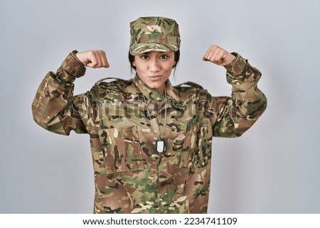 Young south asian woman wearing camouflage army uniform showing arms muscles smiling proud. fitness concept. 