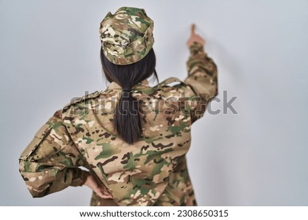 Young south asian woman wearing camouflage army uniform posing backwards pointing ahead with finger hand 