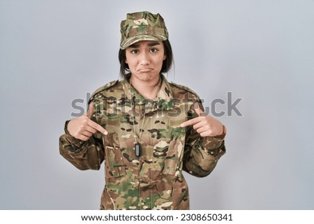 Young south asian woman wearing camouflage army uniform pointing down looking sad and upset, indicating direction with fingers, unhappy and depressed. 