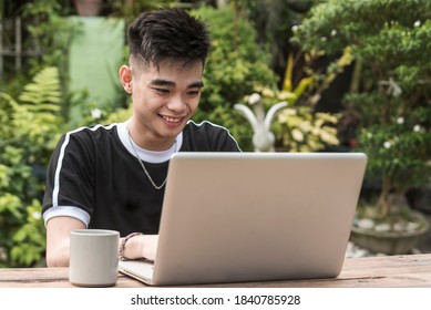 A Young Southeast Asian Man Happily Works On His Laptop While Outside At His Home Or At A Park.