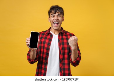 Young Spanish Latinos Man In Red Checkered Shirt Hold Mobile Cell Phone Blank Screen Workspace Show Thumb Up Gesture Area Do Winner Gesture Clench Fist Isolated On Yellow Background Studio Portrait