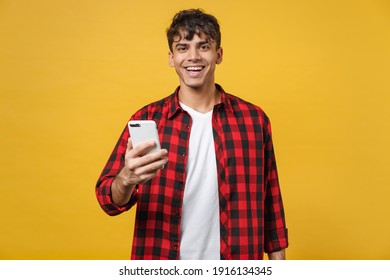 Young Spanish Latinos Smiling Handsome Fashionable Man 20s Wearing Red Checkered Shirt Holding Mobile Cell Phone Chatting With Friends In Social Network Isolated On Yellow Background Studio Portrait