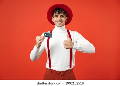 Young Spanish Latinos Smiling Happy Rich Stylish Man 20s In Hat White Shirt Trousers With Suspenders Hold In Hand Credit Bank Card Show Thumb Up Like Gesture Isolated On Red Background Studio Portrait