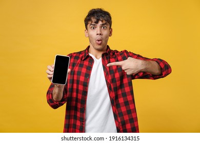 Young Spanish Latinos Surprised Man In Red Checkered Shirt Hold Point Index Finger On Mobile Cell Phone Blank Screen Workspace Show Thumb Up Gesture Area Isolated On Yellow Background Studio Portrait