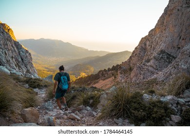 Young Spanish Man Walking Through A Hiking Path In Serra De Tramuntana At Golden Hour With The Village Of Soller At The Backgorund Tinted By Golden Light