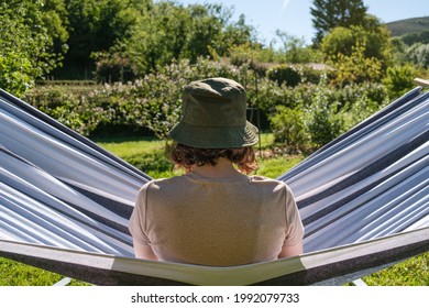 Young Spanish Woman Sitting In Hammock With Her Back Turned Looking At The Garden. She Is Wearing A Bucket Hat.