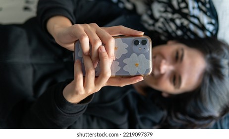 Young Spanish Woman Using The Phone While She Is Lying Down The Sofa And Smiling To It, Focus On Foreground