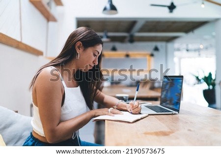 Young Spanish woman work online on laptop computer sitting in coworking, write notes in notebook.Smiling female freelancer browsing website on netbook. Student learning on webinar, reading information