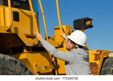 Young Specialist Is Getting Up To Yellow Forklift Operator Cabine Holding Handrails. Safety Glasses And White Hard Hat. 