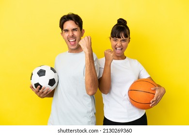 Young Sport Couple Playing Football And Basketball Isolated On Yellow Background Celebrating A Victory