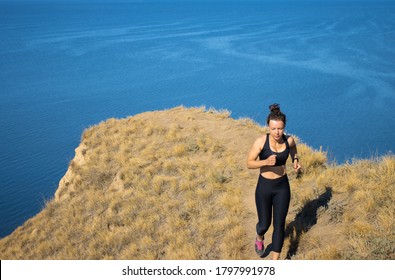 Young Sport Woman In Black Sportswear Trail Running Training Cardio On Rocky Mountain Path Near Blue Sea In Sunny Day. Long Distance Endurance Run In Summer Outdoors Nature Landscape. Copy Space