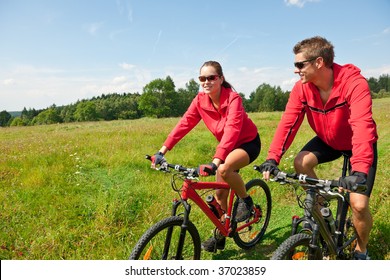 Young Sportive Couple Riding Mountain Bike In Spring Meadow On A Sunny Day