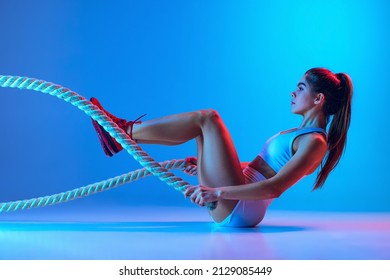 Young Sportive Girl Training With Sports Equipment Isolated On Gradient Blue-pink Studio Background In Neon Light. Modern Sport, Action, Motion, Youth Concept. Fitness, Hobby, Healthy Lifestyle