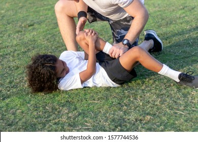 Young Sports Boy With Injured Knee Getting Help From Man Touching His Knee.Injured Boy Lying On The Grass In Pain,making Face Expression.Shot Of Man Looking At Injured Boy And Trying To Help Him.