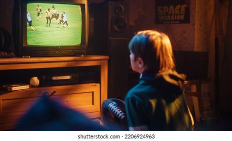 Young Sports Fan Watches American Football Match On TV At Home. Handsome Boy Supporting His Favorite Team. Holding A Football In Excitement. Nostalgic Retro Childhood Concept.