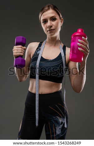 Young sports girl with long blonde hair stuffed in tail, beautiful appearance, sports body, in black top and legines, holds sports water bottle, phone and inch tape around her neck