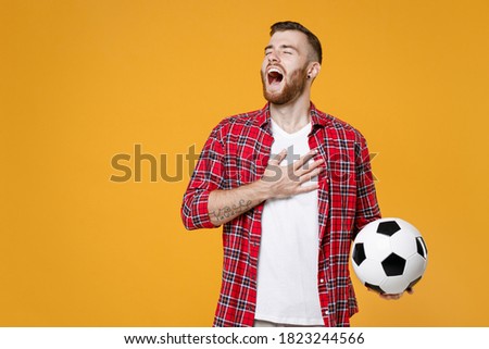 Young sports man football fan in basic red shirt cheer up support favorite team with soccer ball put hand on heart sing anthem isolated on yellow background studio. Tattoo translation life is fight
