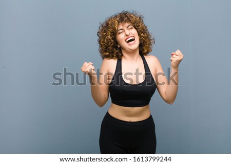young sports woman looking extremely happy and surprised, celebrating success, shouting and jumping against gray wall
