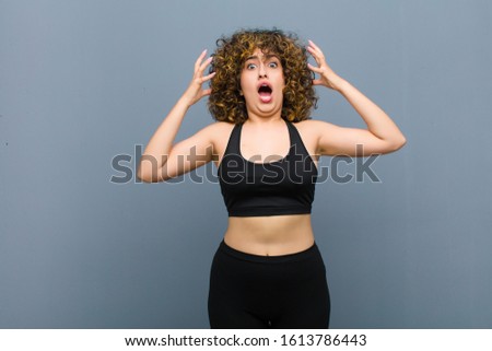 young sports woman screaming with hands up in the air, feeling furious, frustrated, stressed and upset against gray wall