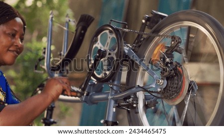 Young sports-loving black woman holding and adjusting bike components in home yard. Enthusiastic female cyclist thoroughly examining and fixing damaged bicycle for summer leisure cycling.