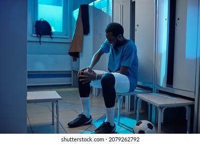 Young Sportsman In Professional Sports Uniform Touching Sick Knee While Sitting By Row Of Lockers In Changing Room