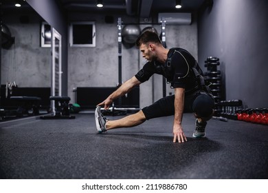 Ein junger Sportler in einem speziellen EMS-Anzug streckt seine Beine aus und erwärmt seine Muskeln, um sich im Fitnessraum zu trainieren. Fitness in EMS-Anzug in einem modernen Fitnesskonzept, revolutionäres Training, elektrische Stimulation