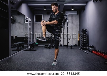 A young sportsman in a special EMS suit stretches and warms up his muscles for training indoor gym. Fitness in EMS suit in a modern gym concept, revolutionary training and care, electrical stimulation
