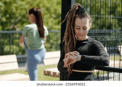 Young sportswoman in black tracksuit looking at smartwatch on her wrist while standing in front of camera against active girl doing exercise - Powered by Shutterstock