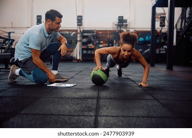 Young sportswoman with one hand on a ball doing push-ups during exercise class with a coach in health club.  - Powered by Shutterstock