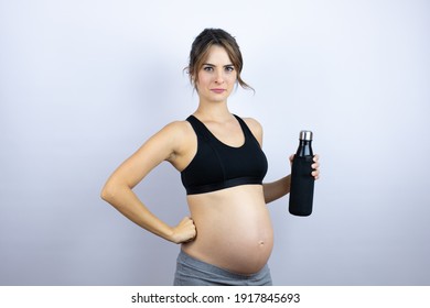 Young sportswoman pregnant wearing sportswear holding bottle with water over white background skeptic and nervous, disapproving expression on face with arms in waist