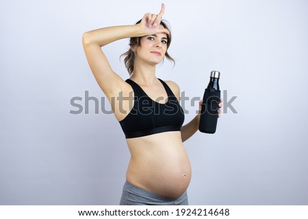 Young sportswoman pregnant wearing sportswear holding bottle with water over white background making fun of people with fingers on forehead doing loser gesture mocking and insulting.