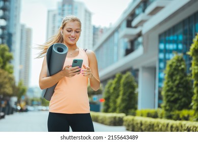 Young sportswoman texting on the street. Sporty, athletic woman listening to music over the wireless earphones in a city street. She has a mobile phone, a yoga mat and a water bottle in her hands. - Powered by Shutterstock