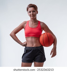 Young sportswoman with toned abs wearing a red sports bra and gray shorts holding a basketball while standing on a white background - Powered by Shutterstock