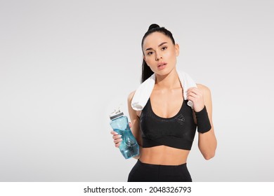 young sportswoman with towel on shoulders holding sports bottle and looking at camera isolated on grey