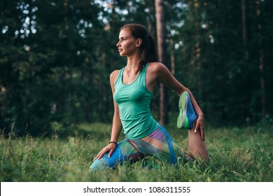 Young sportswoman warming-up before workout doing stretching exercises sitting on grass in park