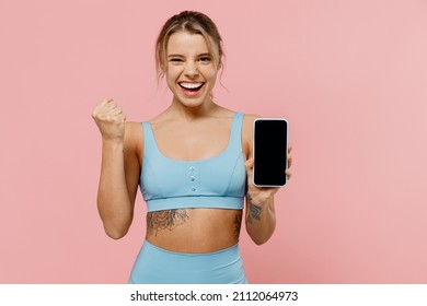 Young sporty athletic fitness trainer instructor woman wear tracksuit spend time in home gym hold mobile cell phone blank screen area do winner gesture isolated on plain pink background Sport concept