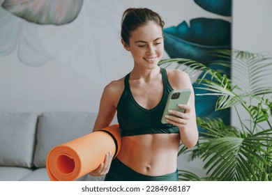 Young sporty athletic fitness trainer instructor woman wearing green tracksuit hold in hand yoga mat use mobile cell phone training do exercises at home gym indoor. Workout sport motivation concept