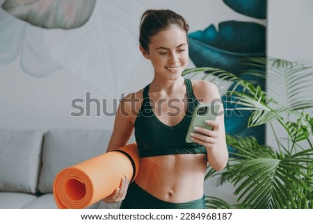 Young sporty athletic fitness trainer instructor woman wearing green tracksuit hold in hand yoga mat use mobile cell phone training do exercises at home gym indoor. Workout sport motivation concept