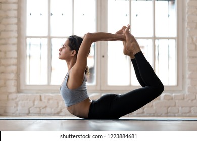 Young sporty attractive woman practicing yoga, doing Dhanurasana exercise, Bow pose, working out, wearing sportswear, pants and top, indoor full length, yoga studio - Shutterstock ID 1223884681
