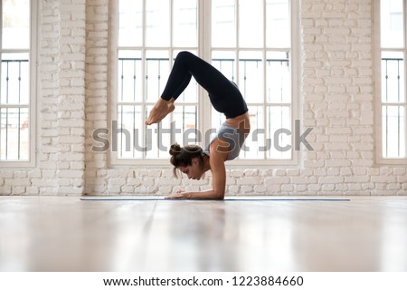 Young sporty attractive woman practicing yoga, doing Vrischikasana, Scorpion exercise, handstand pose, working out, wearing sportswear, pants and top, indoor full length, yoga studio