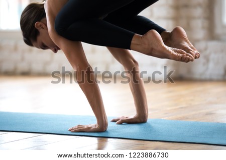 Young sporty attractive woman practicing yoga, doing Crane exercise, Bakasana pose, working out, wearing sportswear, pants and top, indoor close up, white yoga studio. Well being concept