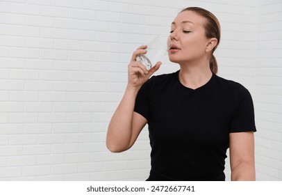 Young sporty female athlete hydrating with a glass of water after a workout in a home studio. The woman is wearing a black shirt and appears refreshed, standing against a white brick background. - Powered by Shutterstock