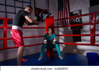 Young sporty female boxer in boxing gloves after battle sitting next coach near red corner of a regular boxing ring in a gym - Powered by Shutterstock