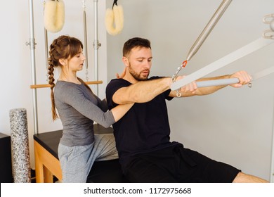 Young Sporty Female Instructor Of Pilates Helping Beard Man Workout In Cadillac Bed. Two People Working In Pilates Studio, Woman Assistant Supporting And Correcting Male Patient Beginner.