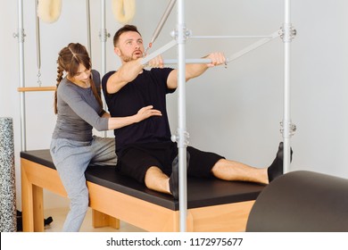 Young Sporty Female Instructor Of Pilates Helping Beard Man Workout In Cadillac Bed. Two People Working In Pilates Studio, Woman Assistant Supporting And Correcting Male Patient Beginner.